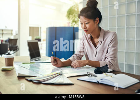 Jeune designer asiatique assis seul à un bureau dans un bureau à la recherche sur la conception des immeubles et de travailler sur un ordinateur portable Banque D'Images
