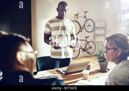 Jeune homme africain donnant une présentation aux collègues assis ensemble autour d'une table dans un bureau moderne Banque D'Images