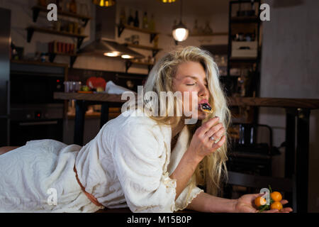 Cute blonde en peignoir se trouve sur la table contre cuisine moderne. Jeune femme heureuse mange des pancakes aux fruits sur le petit-déjeuner à home Banque D'Images