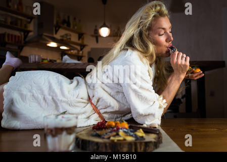 Cute blonde en peignoir se trouve sur le tableau à l'élégant dans le matin. Jeune femme heureuse mange des pancakes sur le petit-déjeuner à home Banque D'Images