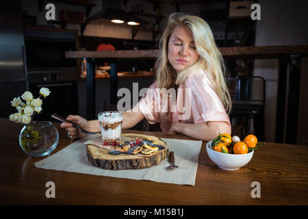 Jeune femme mange le petit-déjeuner dans le salon. Blonde pensif assis à la table avec la nourriture contre cuisine moderne au petit matin Banque D'Images