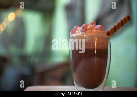 Délicieux chocolat chaud boire dans un verre garni de crème fouettée guimauves rose avec un bâton de chocolat et de flou arrière-plan de la salle en vert clair Banque D'Images