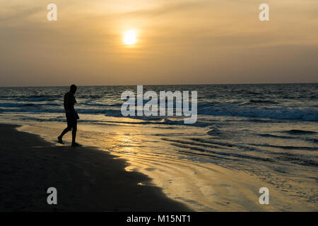 Un homme africain se profile comme il marche le long du rivage de Kotu Beach en Gambie au coucher du soleil. Banque D'Images