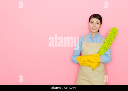 Heureux confiant service propre entreprise employée portant des gants et tablier avec plumeau comptable permanent de l'outil à fond rose. Banque D'Images