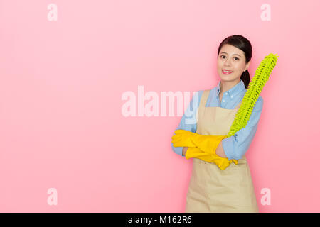 Certains jeunes femmes houseworker holding Feather Duster debout dans l'outil de nettoyage de fond rose et portant des gants jaunes looking at camera smilin Banque D'Images