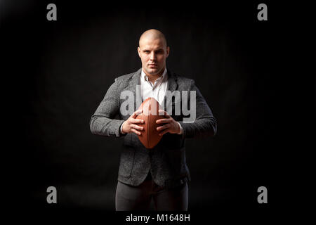 Un jeune homme chauve dans un costume gris, chemise blanche tient un ballon de rugby sur un fond isolé noir Banque D'Images