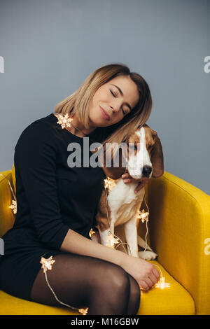 Jeune femme en robe noire hugs beagle puppy sur chaise jaune sur fond de mur gris avec Banque D'Images