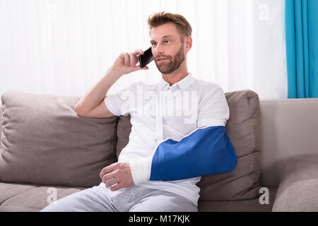 Jeune homme avec la main fracturée Sitting on Sofa Talking On Mobile Phone Banque D'Images