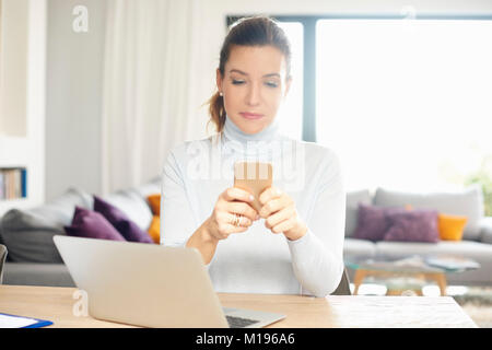 Jolie femme en utilisant son téléphone mobile et de messagerie texte tout en travaillant sur l'ordinateur portable de la maison. Banque D'Images