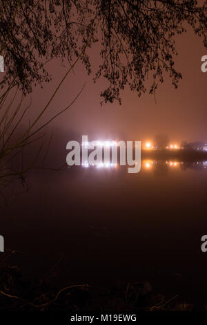 Le brouillard au-dessus d'un lac ou un étang dans la nuit. La vue à l'horizon, les lumières de la ville et des bâtiments dans l'obscurité. Encadrée par de sombres silhouettes d'un arbre. Banque D'Images