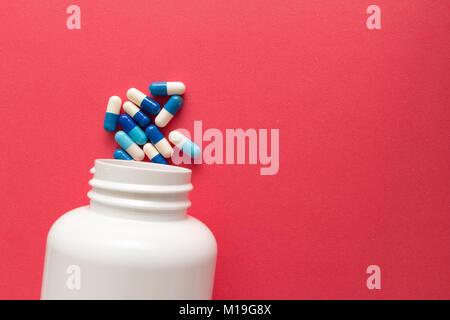 Groupe d'autres comprimés blanc et bleu. Capsules spilling out of white bouteille. Fond rouge. Banque D'Images