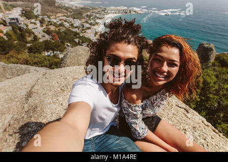 Jeune homme et femme assis sur le haut de falaise taking self portrait. Heureux couple assis sur le sommet de la montagne et en tenant. selfies Banque D'Images