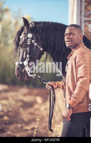 African Man wearing sunglasses près de black horse in hangar Banque D'Images