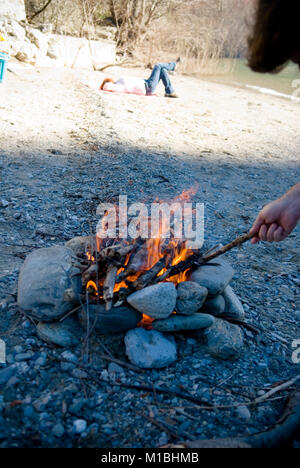 Au premier plan un homme tend à un feu sur une plage au bord d'une rivière alors qu'une femme se trouve avec les jambes croisées à l'arrière-plan Banque D'Images