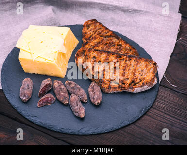 Morceau de fromage et deux toasts de pain de seigle, à côté de morceaux de saucisse salée sur un fond noir, vue du dessus Banque D'Images