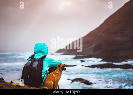 Homme avec un chien en face de paysage littoral rural avec des montagnes et des vagues venant et rayons de soleil à travers les nuages. L'île de Santo Antao, Cap Vert Banque D'Images