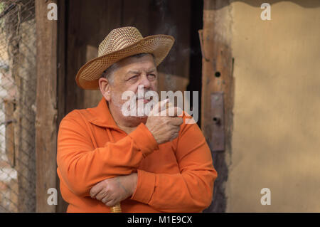 Beau portrait de barbu ukrainien senior man in straw hat assis contre l'ancienne grange avec tuyau d'entrée à l'usage du tabac Banque D'Images