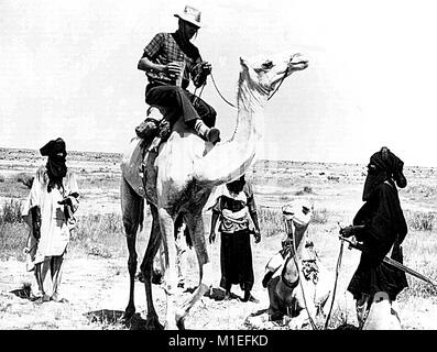 Image en noir et blanc illustrant quatre hommes et deux chameaux, trois hommes en vêtements d'Afrique, l'un tenant la tête d'un chameau, se tenir assis en demi-cercle autour de centres de contrôle des maladies (CDC) Operations Officer Anthony R. Masso qui monte un chameau dans le Niger, l'Afrique, au cours d'une épidémie de variole, 1975. Image courtoisie CDC. () Banque D'Images