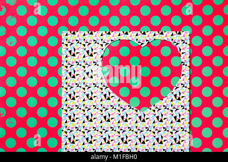 Un cœur découpé sur un papier à motifs abstraits colorés, et un fond rouge à motifs de points verts vu à travers l'espace découpé Banque D'Images