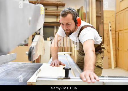 Friendly carpenter avec protège-oreilles et des vêtements de travail travaillant sur une scie dans l'atelier Banque D'Images