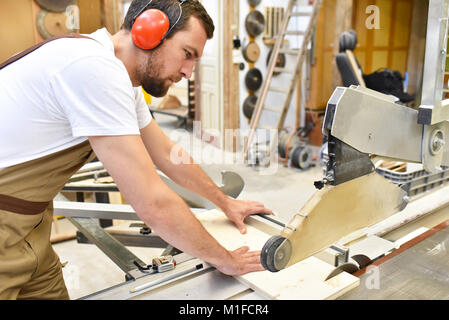 Friendly carpenter avec protège-oreilles et des vêtements de travail travaillant sur une scie dans l'atelier Banque D'Images