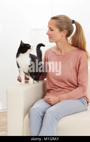 Chat domestique. Les jeunes et adultes de smoking femme assise sur un canapé blanc. Allemagne Banque D'Images