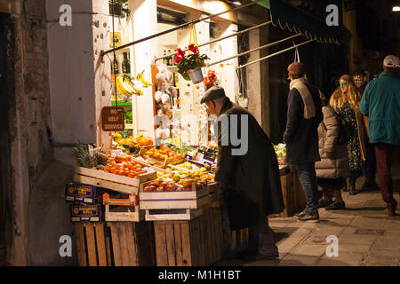 Vieil homme shopping pour les fruits de nuit à Cannaregio, Venise, Italie à un magasin de la rue en plein air avec un jeune couple de carnaval en passant b Banque D'Images