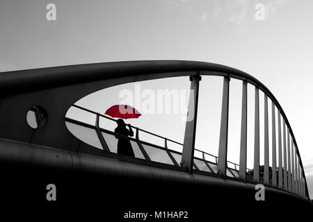 Une photo d'une dame debout sur un pont avec un parapluie rouge au cours de la jour de pluie. Le parapluie est isolé dans l'image en noir et blanc. Banque D'Images