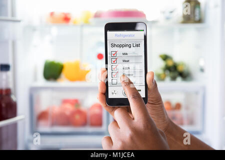 Close-up de la main de l'homme marquant la liste d'achats sur téléphone mobile devant ouvrir un réfrigérateur Banque D'Images