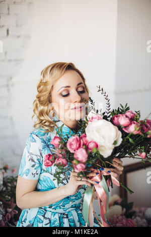 Femme blonde en robe bleu clair avec des fleurs en choisissant l'avant du mur de lumière et l'odeur des fleurs Banque D'Images