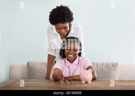 Une petite fille africaine assis avec sa mère le passage des pièces de Pink Piggy Bank à la maison Banque D'Images