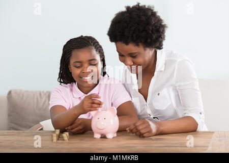 Une petite fille africaine assis avec sa mère le passage des pièces de Pink Piggy Bank à la maison Banque D'Images