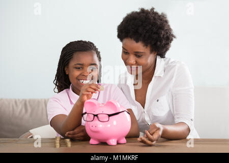 Une petite fille africaine assis avec sa mère le passage des pièces de Pink Piggy Bank à la maison Banque D'Images