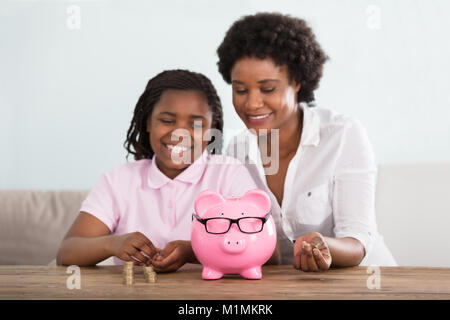 Une petite fille africaine assis avec sa mère le passage des pièces de Pink Piggy Bank à la maison Banque D'Images