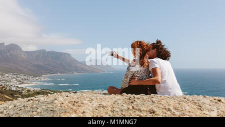 Couple assis sur le sommet de la montagne et en tenant. selfies Jeune homme et femme assis ensemble à l'extérieur sur falaise taking self portrait with mobile phone Banque D'Images