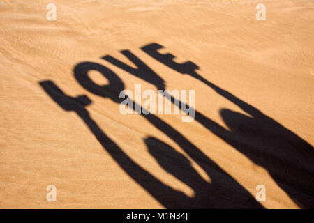 Gros plan de l'ombre d'un homme et une femme tenant quelques lettres formant le mot amour au-dessus de leur tête sur le sable d'une plage Banque D'Images