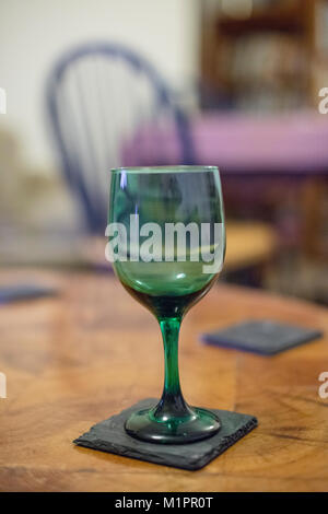 Un verre de vin vert sur un coaster en ardoise sur une table basse en bois avec une chaise et une table à manger dans l'arrière-plan Banque D'Images