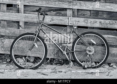 Un vélo abandonné appuyé contre une barrière en bois. La photo est en noir et blanc. Banque D'Images