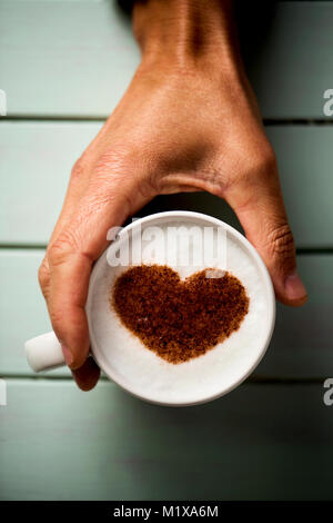 Capture d'un grand angle d'un jeune homme de race blanche avec une tasse de Cappuccino avec un cœur dessiné avec du cacao en poudre sur sa mousse de lait, d'un vert pâle en bois rustique Banque D'Images