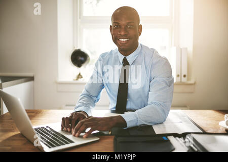 Jeunes hommes assis à son bureau dans un bureau de sourire et de travailler sur un ordinateur portable Banque D'Images