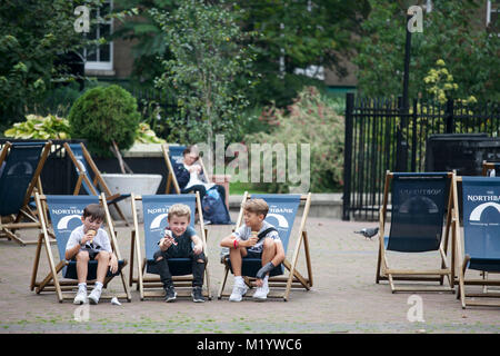Londres, ANGLETERRE - 22 août 2017 Trois garçons de 6 ans sont la consommation de crème glacée assis sur des chaises longues dans le parc Banque D'Images
