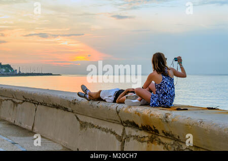 Jeune couple assis, couché sur le bord de l'eau regarder et photographier le coucher du soleil sur l'océan Banque D'Images