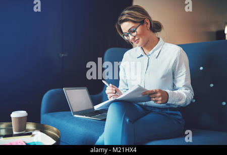 Smiling young businesswoman reading à travers des documents et à l'aide d'un ordinateur portable tout en étant assis sur un canapé à la maison Banque D'Images