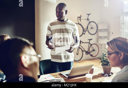 L'accent young African man parlant à un groupe de collègues assis ensemble autour d'une table dans un bureau moderne Banque D'Images