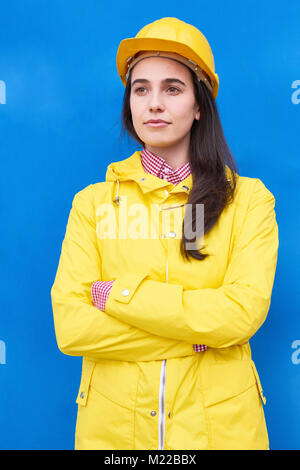 Bright colorful portrait of young woman wearing hardhat et yellow jacket article contre mur bleu contrasté posing with arms crossed Banque D'Images
