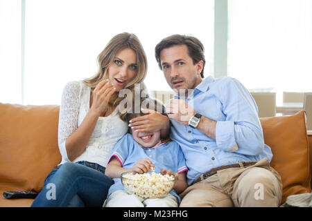 Beaux jeunes parents et leurs fils sont devant la télé, fermer les yeux du garçon Banque D'Images