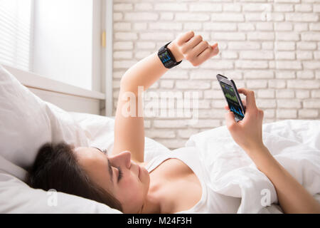 Close-up of a Young Woman Lying On Bed Synchronisation Smart Watch With Cell Phone Banque D'Images