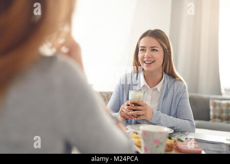 Happy smiling adolescent ayant le petit déjeuner à la maison assis à la table avec une boisson et de la transmission à la personne en face qui est hors de l'écran Banque D'Images