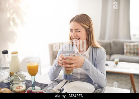 Happy smiling adolescent ayant le petit déjeuner à la maison assis à la table avec une boisson et de la transmission à la personne en face qui est hors de l'écran Banque D'Images