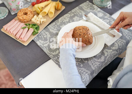 Woman slicing un rouleau du pain sur une plaque à la table avec un assortiment de charcuterie et fromage sur un plat en face d'elle Banque D'Images